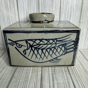 May include: A rectangular ceramic container with a fish design. The container is light beige with a dark blue fish illustration on the front. The top has a small, round opening. The container is sitting on a wooden surface.