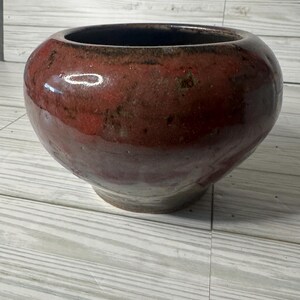 May include: A round, glazed ceramic bowl with a wide opening. The bowl is a deep red with darker accents near the rim and a lighter base. The interior is a dark brown. The bowl sits on a light-coloured wooden surface.