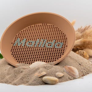Könnte beinhalten: Ein braunes Plastiksieb mit einem Gittermuster und dem aufgedruckten Wort "Matilda". Das Sieb liegt auf einem Sandhaufen mit Muscheln.