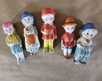 May include: Five porcelain dolls with painted faces and clothing. The dolls are wearing various outfits, including a red dress, a blue dress, a red and white outfit, a blue and white outfit, and a blue and white outfit. The dolls are all standing on a brown surface.