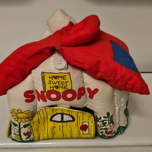 May include: A plush Snoopy dog house toy with a red roof and a yellow door. The house has the words "Home Sweet Home" on the front and the word "Snoopy" in large letters.