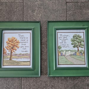 May include: Two green picture frames with white mats and embroidered images. The left frame has a yellow tree and the text "Dad, there is never a moment that you are not loved." The right frame has a green tree and the text "Mother, you give so much and ask so little in return."