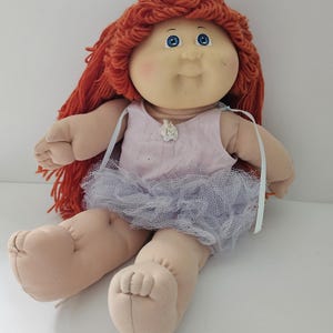 May include: A vintage doll with a soft, fabric body and a vinyl face. The doll has bright blue eyes, rosy cheeks, and red yarn hair. It wears a pink dress with a light blue tulle skirt. A classic toy.