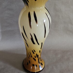 May include: A tall, cream-colored glass vase with a brown and black animal print design. The vase has a wide mouth and a narrow base.