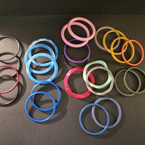 May include: A collection of 15 colorful plastic bangles in various shades of blue, green, purple, pink, red, and yellow. The bangles are stacked and arranged on a black surface.