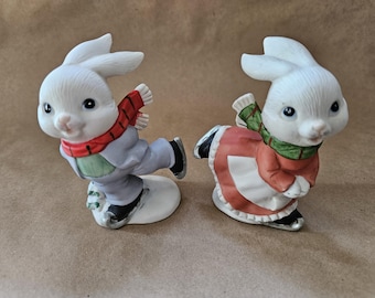 May include: Two white porcelain bunny figurines ice skating. The boy bunny is wearing a blue and green outfit with a red scarf. The girl bunny is wearing a red and green dress with a green scarf.