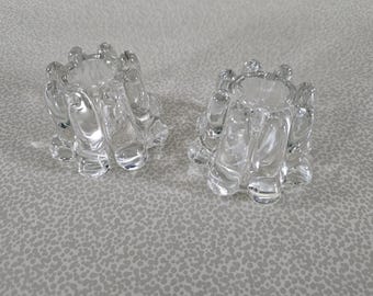May include: Two clear glass candle holders with a unique, organic design. Each holder has a circular opening surrounded by curved, bubble-like shapes. The glass is transparent, allowing light to pass through. The candle holders are placed on a light gray surface.