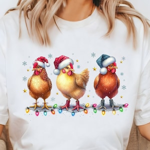 Puede incluir: Camiseta blanca con un gráfico de tres pollos con gorros de Papá Noel y de pie frente a una cadena de luces navideñas.