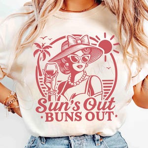 May include: A white t-shirt with a red graphic of a woman wearing a hat and sunglasses holding a glass of wine. The graphic also includes a palm tree, a sun, and the text "Sun's Out Buns Out".