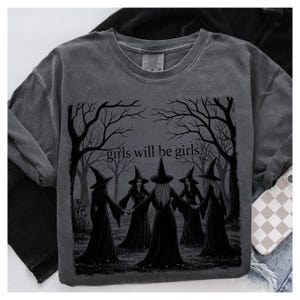 K&ouml;nnte beinhalten: Dunkelgraues Sweatshirt mit einer Grafik von f&uuml;nf Hexen, die sich im Kreis an den H&auml;nden halten, umgeben von kahlen B&auml;umen. Der Text "girls will be girls" steht &uuml;ber den Hexen. Das Sweatshirt hat einen Rundhalsausschnitt und lange &Auml;rmel.