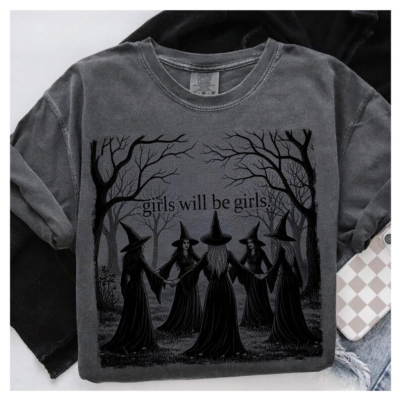 K&ouml;nnte beinhalten: Dunkelgraues Sweatshirt mit einer Grafik von f&uuml;nf Hexen, die sich im Kreis an den H&auml;nden halten, umgeben von kahlen B&auml;umen. Der Text "girls will be girls" steht &uuml;ber den Hexen. Das Sweatshirt hat einen Rundhalsausschnitt und lange &Auml;rmel.