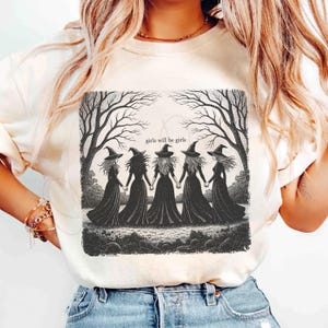 May include: Cream-colored t-shirt featuring a black and white illustration of five witches holding hands under bare trees. The text "girls will be girls" is above the witches. The shirt has short sleeves.