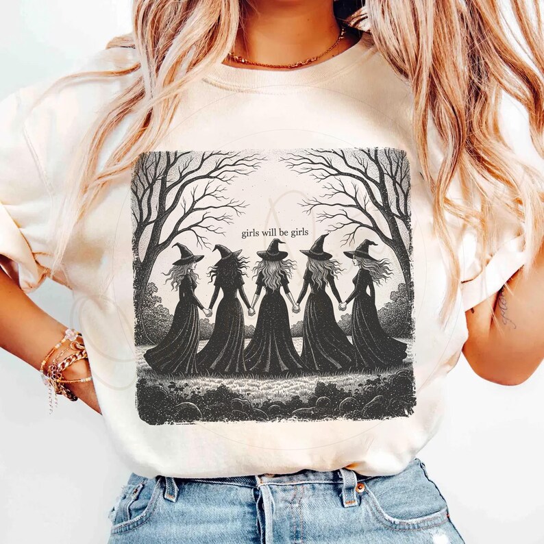 May include: Cream-colored t-shirt featuring a black and white illustration of five witches holding hands under bare trees. The text "girls will be girls" is above the witches. The shirt has short sleeves.