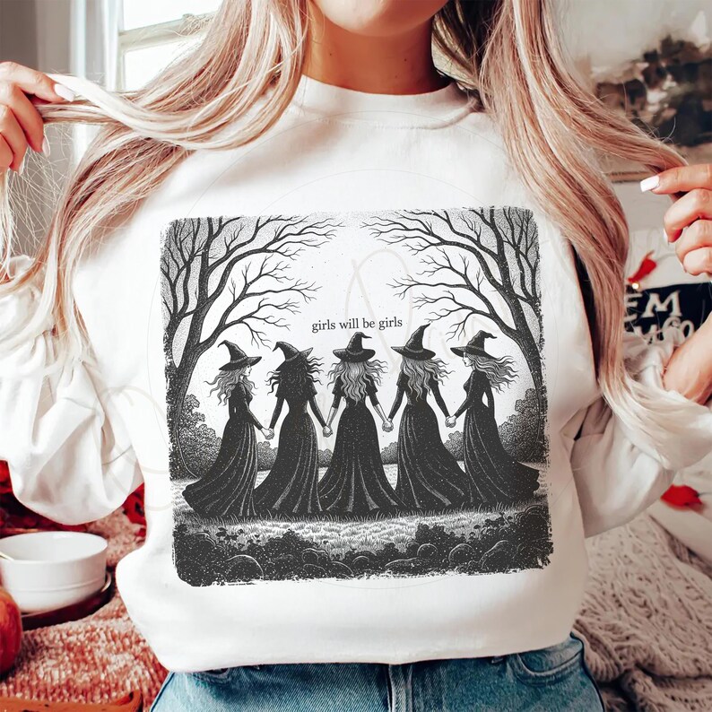 May include: White sweatshirt featuring a black and white illustration of five witches holding hands. The witches are wearing long black dresses and pointed hats. The text "girls will be girls" is above the witches.