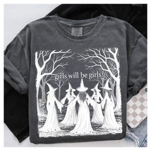 K&ouml;nnte beinhalten: Dunkelgraues T-Shirt mit einer wei&szlig;en Grafik von f&uuml;nf Figuren mit Hexenh&uuml;ten, die sich an den H&auml;nden halten, umgeben von B&auml;umen. Der Text "girls will be girls" befindet sich dar&uuml;ber.