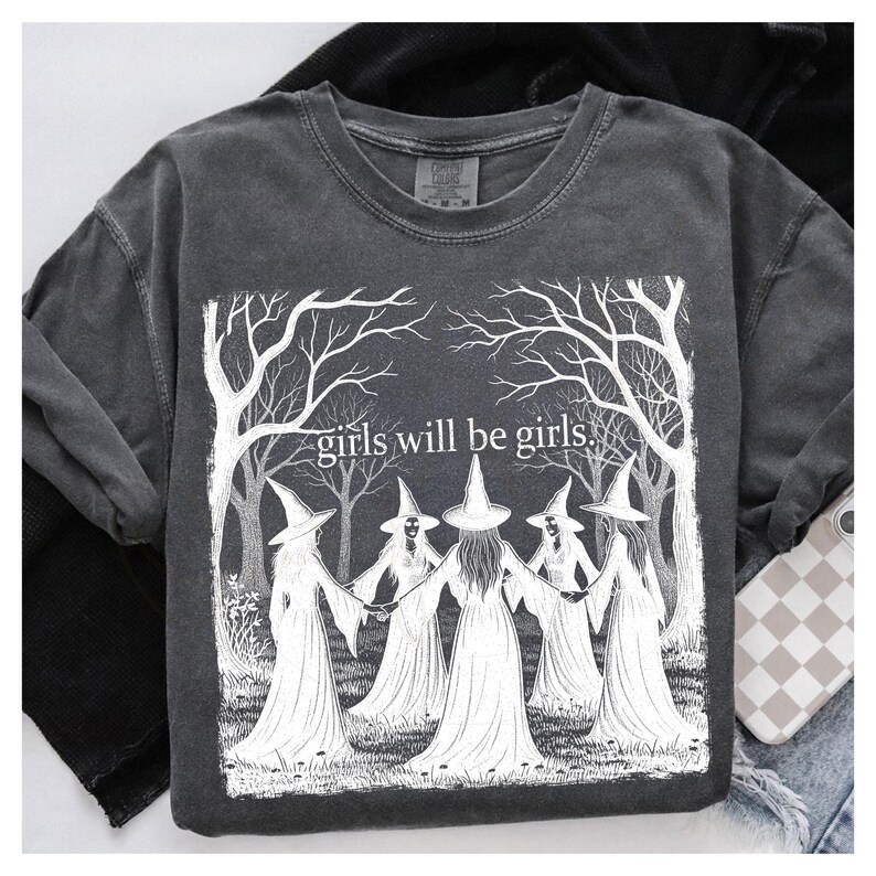 K&ouml;nnte beinhalten: Dunkelgraues T-Shirt mit einer wei&szlig;en Grafik von f&uuml;nf Figuren mit Hexenh&uuml;ten, die sich an den H&auml;nden halten, umgeben von B&auml;umen. Der Text "girls will be girls" befindet sich dar&uuml;ber.
