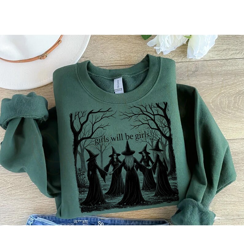 K&ouml;nnte beinhalten: Dunkelgr&uuml;nes Sweatshirt mit einem schwarzen Aufdruck von f&uuml;nf Hexen mit spitzen H&uuml;ten, die sich in einem Wald an den H&auml;nden halten. Der Text "girls will be girls" steht &uuml;ber den Hexen. Rundhalsausschnitt und lange &Auml;rmel.