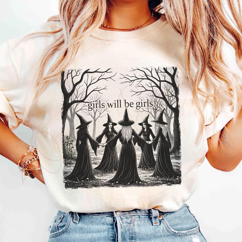 K&ouml;nnte beinhalten: Cremefarbenes Sweatshirt mit einer Schwarz-Wei&szlig;-Illustration von f&uuml;nf Hexen in schwarzen Roben und H&uuml;ten, die sich an den H&auml;nden halten. Der Text "girls will be girls" befindet sich &uuml;ber der Illustration, vor einem Waldhintergrund.