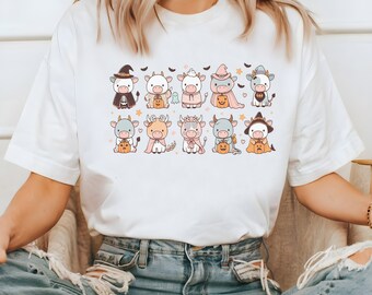 May include: White t-shirt with a graphic of ten cartoon cows dressed in Halloween costumes. The cows are wearing hats, costumes, and holding pumpkins.