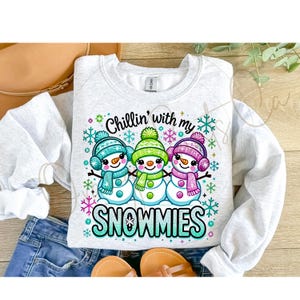 May include: A white sweatshirt with a graphic of three snowmen holding hands. The snowmen are wearing colorful hats, scarves, and earmuffs. The text "Chillin' with my Snowmies" is printed in turquoise glitter letters.