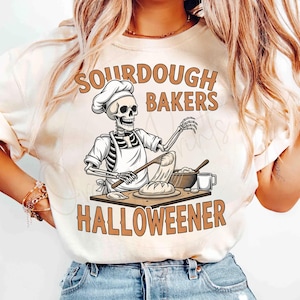 Könnte beinhalten: Beiges Sweatshirt mit Rundhalsausschnitt und Halloween-Motiv. Das Motiv zeigt einen Skelettbäcker mit Kochmütze und Schürze, mit den Worten "SOURDOUGH BAKERS HALLOWEENER" in braunen Buchstaben.