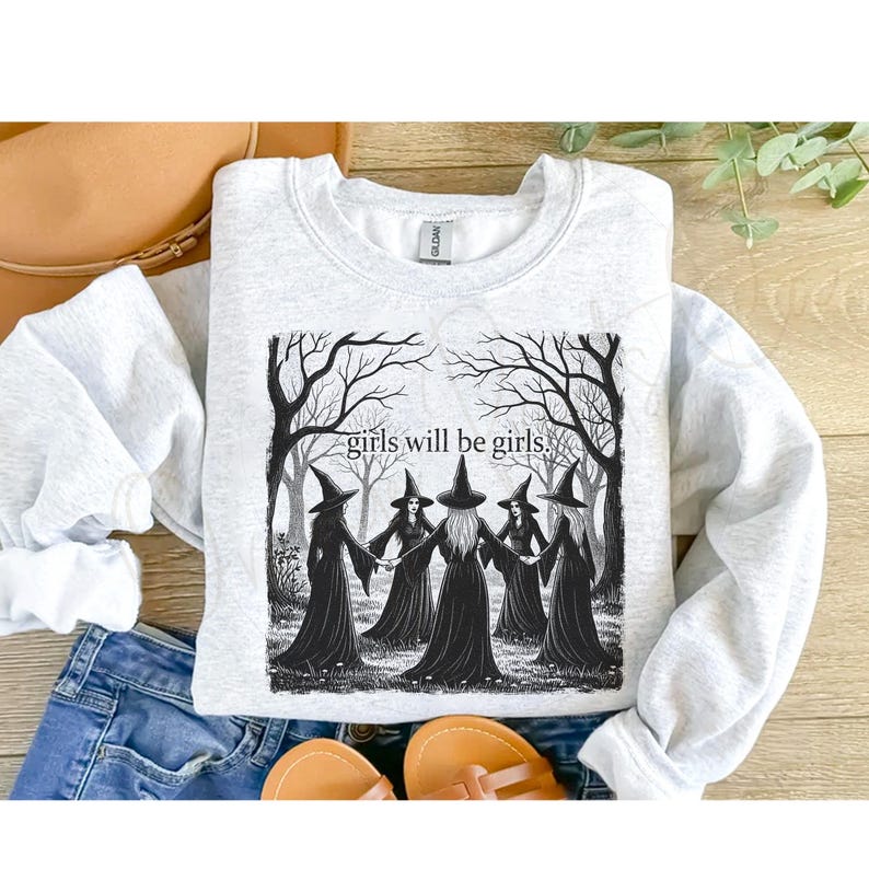 K&ouml;nnte beinhalten: Ein hellgraues Sweatshirt mit einer Schwarz-Wei&szlig;-Grafik von sechs Hexen in einem Kreis, die sich unter kahlen B&auml;umen an den H&auml;nden halten. Der Text "girls will be girls" steht &uuml;ber den Hexen. Das Sweatshirt liegt auf einer Holzoberfl&auml;che mit Hut und Jeans.