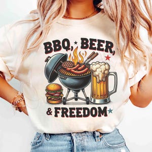 May include: White t-shirt with a graphic design of a grill, beer, and burger. The text on the shirt reads "BBQ, BEER, & FREEDOM".
