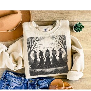 May include: A beige sweatshirt with a graphic of five witches holding hands under bare trees, with the text "girls will be girls". The sweatshirt is paired with blue jeans, a brown hat, and sandals.