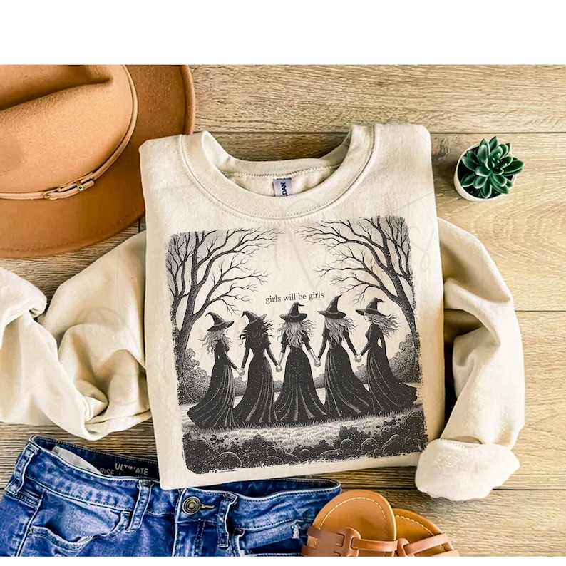 May include: A beige sweatshirt with a graphic of five witches holding hands under bare trees, with the text "girls will be girls". The sweatshirt is paired with blue jeans, a brown hat, and sandals.