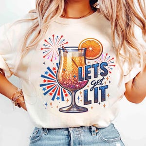以下が含まれることがあります： クリーム色のTシャツ。カクテルのデザインと「Lets Get Lit」の文字。赤、白、青のアクセントで、オレンジスライス、花火、星が描かれたカクテルグラスのグラフィック。