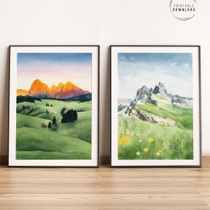 Puede incluir: Dos impresiones enmarcadas de acuarela de paisajes de montaña. Una presenta montañas doradas al atardecer sobre colinas verdes con una pequeña cabaña. La otra representa una montaña rocosa con prados verdes y flores amarillas.