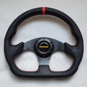 May include: Black steering wheel with red stitching and a red stripe at the top. The center features a black hub with the "momo" logo in yellow. The wheel has a sporty design.