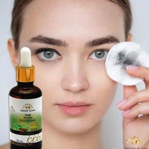 May include: A 50 ml bottle of Desert Elixir Jojoba Oil with a gold cap and label. The label reads "Desert Elixir, Huile de Jojoba, Démaquillant Naturel, 100% Pure, Jojoba Oil". A cotton pad with black makeup residue is being used to remove makeup from a person's eye.