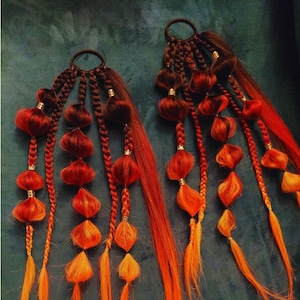 Puede incluir: Dos juegos de extensiones de cabello con múltiples trenzas. Cada trenza tiene pequeñas cuentas rojas, naranjas y amarillas. Las trenzas están unidas a una liga para el cabello negra.