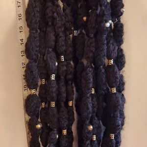 Può includere: Due extension per dreadlock nere con dettagli oro e argento. I dreadlock sono adornati con piccole perline dorate e perle argentate. Le extension sono appese verticalmente, mostrando la loro lunghezza e consistenza.