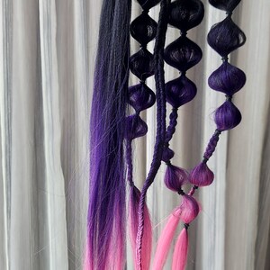 Può includere: Un'estensione per capelli lunga, nera, viola e rosa con più trecce e piccoli elastici rotondi viola.