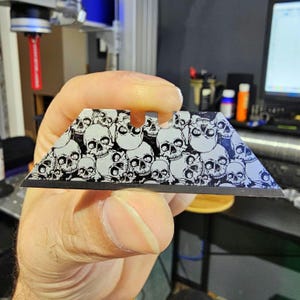 May include: A silver utility knife blade with a black skull pattern. The blade is designed for use in a utility knife.