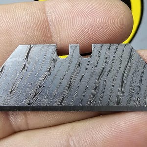 May include: A silver utility knife blade with a patterned finish.