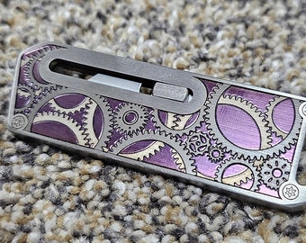 May include: A small, silver-coloured pocket knife with a purple and gold gear pattern. The knife has a blade that slides out of the side.