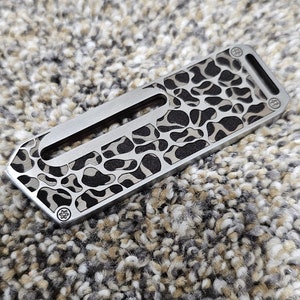May include: A rectangular, stainless steel pocket knife with a unique pattern. The knife features a series of irregular shapes cut into the metal, creating a textured appearance. The design is a mix of silver and black.