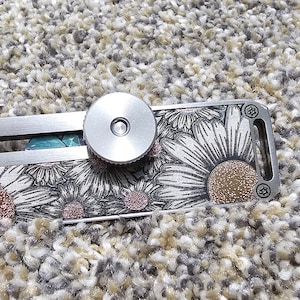 May include: A silver metal keychain with a floral design and a rotating dial. The design features white daisies with pink centers on a black background.