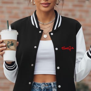May include: Black and white letterman jacket with the word "SUGA" in red script on the chest. The jacket has white sleeves and striped cuffs. The person is wearing a white crop top and holding a coffee cup.