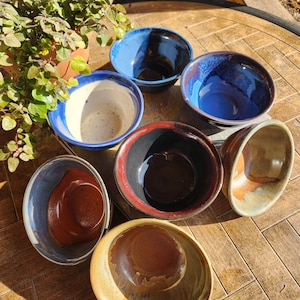 May include: A collection of six ceramic bowls in various colors and patterns. The bowls are arranged on a wooden surface. The bowls are all different sizes and shapes.