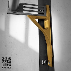 K&ouml;nnte beinhalten: Eine goldfarbene Metallhalterung, montiert an einer schwarzen vertikalen St&uuml;tze, mit einer passenden horizontalen St&uuml;tze. Die Halterung hat ein dreieckiges Design mit einem gebogenen unteren Abschnitt. Das Bild ist vor einem neutralen Hintergrund.