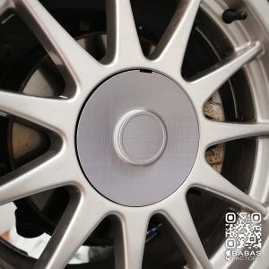 May include: A close-up shot of a silver car wheel, featuring a central hubcap. The wheel has multiple spokes extending from the centre. The hubcap has a brushed metal finish with a circular indentation. A QR code is visible in the bottom right corner.