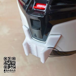 May include: Close-up of a white and black helmet with a red "LOCK" button. A white 3D-printed component is attached to the helmet, with the text "BABAS FACTORY" and a QR code visible. A black cord is attached to the white component.