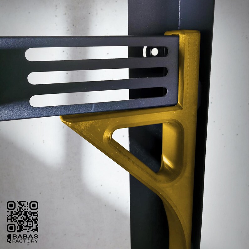 K&ouml;nnte beinhalten: Nahaufnahme einer schwarzen Metallhalterung mit drei horizontalen Schlitzen, verbunden mit einer goldfarbenen, dreieckigen St&uuml;tzhalterung. Die St&uuml;tzhalterung hat ein Aussparungsdesign. Das Bild enth&auml;lt einen QR-Code und den Text "BABAS FACTORY".