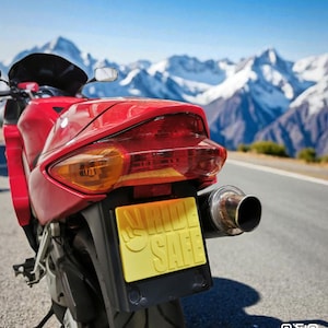 May include: A red motorcycle with a yellow license plate that reads "RIDE SAFE." The motorcycle is parked on a road with a mountain range in the background. The exhaust pipe is silver.