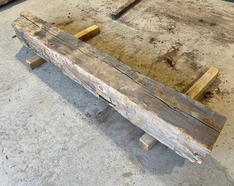May include: A weathered, rectangular wooden beam, likely reclaimed, with a rough, textured surface and visible cracks. The beam is supported by four small, light-colored wooden blocks. The wood has a natural, aged appearance, with shades of gray and brown.
