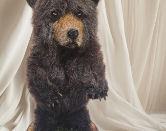May include: A handcrafted, standing bear figurine with dark brown fur and a lighter brown snout. The bear is posed on a wooden base adorned with green moss and small yellow flowers. The background is a soft, off-white fabric.